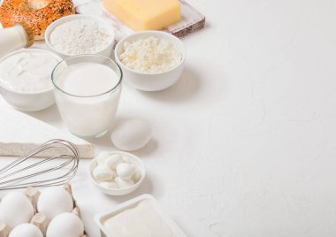 Beyaz masa arka planında taze süt ürünleri. Bir bardak süt, bir kase ekşi krema, süzme peynir ve yumurta. Taze pişmiş simit. Çelik çırpıcı. Üst görünüm.