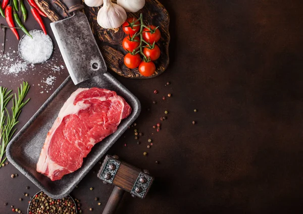 Bife de vaca lombo cru em bandeja de plástico com sal e pimenta e incubatórios de carne vintage e martelo em fundo enferrujado. Pimenta vermelha, tomates e alho. Espaço para texto — Fotografia de Stock