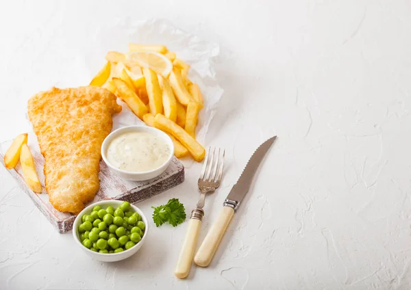 Tradiční britské ryby a hranolky s tatarkou na prkénko s vidličkou a nožem a zelený hrášek na bílé kamenné pozadí stolu. Prostor pro text — Stock fotografie