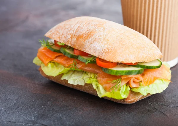 Φρέσκο υγιεινό σολομού σάντουιτς με μαρούλι και αγγούρι με χαρτί κύπελλο καφέ σε μαύρο φόντο πέτρα. Πρωινό σνακ. — Φωτογραφία Αρχείου