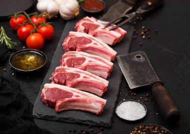 Taze çiğ kasaplar vintage et çatalı ve bıçağı ve balta siyah arka plan üzerine taş tahta üzerinde dana pirzola kuzu. Tuz, biber ve yağ ile domates ve sarımsak ve Barbekü sos.