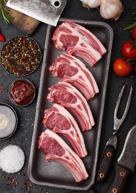 Taze çiğ kasapların vintage et balta ile plastik tepsi ve çatal bıçak siyah arka plan üzerine ile dana pirzola kuzu. Tuz, biber, domates ve sarımsak ve petrol.