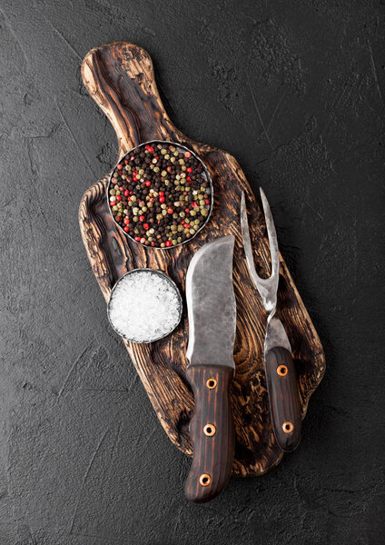 Vintage meat knife and fork on vintage chopping board and black stone table background. Butcher utensils. Salt and pepper.