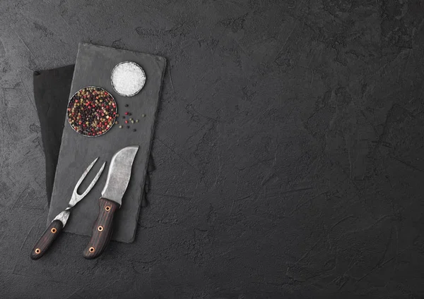 Vintage maso nůž a vidličku s kamenem prkénko a černý stůl pozadí. Řezník nádobí. Sůl a pepř. — Stock fotografie