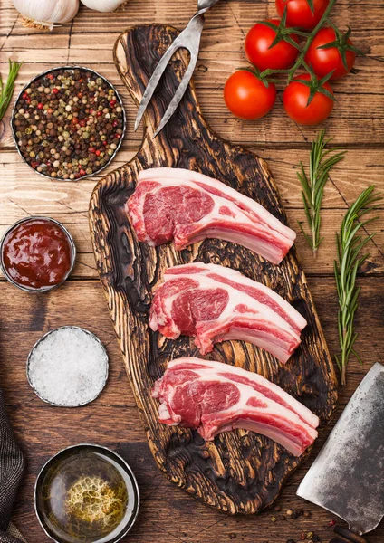 Carniceiros crus frescos costeletas de carne de cordeiro em tábua de corte com hatchets de carne vintage em fundo de madeira.Sal, pimenta e óleo com tomate e alho e molho e garfo de churrasco — Fotografia de Stock