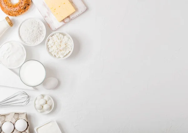 Fresh dairy products on white table background. Glass of milk, bowl of sour cream and cottage cheese and eggs. Fresh baked bagel. Steel whisk. Top view.Space for text