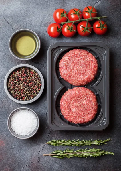 Plastic tray met rauwe gehakte zelfgemaakte vlees rundvlees hamburgers met kruiden en kruiden. Bovenaanzicht. Op steenkeuken tafel achtergrond met tomaten — Stockfoto