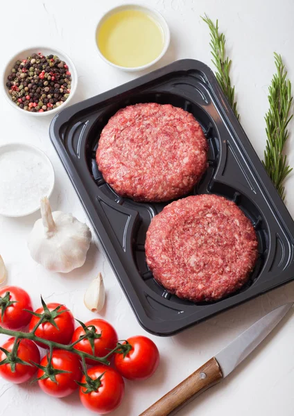 Plastic tray met rauwe gehakte zelfgemaakte rundvlees hamburgers met kruiden en kruiden. Bovenaanzicht. Op de top van de witte keukentafel achtergrond met tomaten zout en peper. — Stockfoto