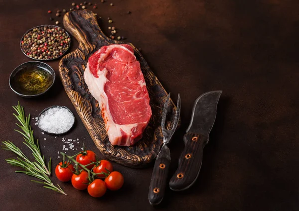 Raw sirloin beef steak on old vintage chopping board with knife and fork on rusty background. Salt and pepper with fresh rosemary and tomatoes.Space for text — Stock Photo, Image