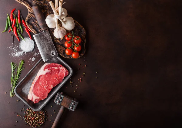 Bife de vaca lombo cru em bandeja de plástico com sal e pimenta e incubatórios de carne vintage e martelo em fundo enferrujado. Pimenta vermelha, tomates e alho. Espaço para texto — Fotografia de Stock