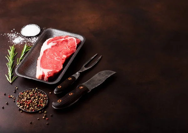 Bife de vaca lombo cru em bandeja de plástico com faca e garfo em fundo enferrujado. Sal e pimenta com alecrim fresco — Fotografia de Stock