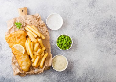 Geleneksel Ingiliz balık ve cips tahta ve beyaz taş masa arka planda yeşil bezelye üzerinde tartar sos ile. limon dilim ve tuz