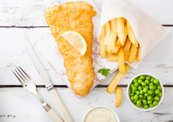 白い木製の背景にフォークとナイフとグリーンピースでまな板の上にタルタルソースを持つ伝統的な英国の魚とチップス. — ストック写真