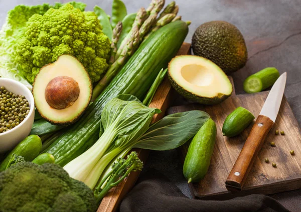 Assorted green toned raw organic vegetables in wooden box on dark background. Avocado, cabbage, cauliflower and cucumber with trimmed beans and chopping board with knife. — Stock Photo, Image