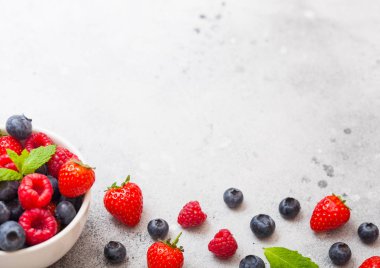 Mutfak masası arka planda beyaz seramik kase plaka taze ham organik çilek. Metin için boşluk. Üst te. Çilek, Ahududu, Yaban Mersini ve Nane yaprağı