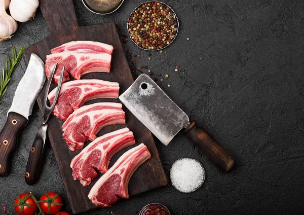 Fresh raw butchers lamb beef cutlets on stone board with vintage meat fork and knife and hatchet on black background.Salt, pepper and oil with tomatoes and garlic and barbeque sauce.Space for text — Stock Photo, Image