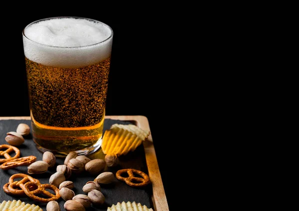Vaso de cerveza lager con merienda sobre tabla de piedra sobre fondo negro. Pistachos y pretzel con patatas fritas. Espacio para texto —  Fotos de Stock