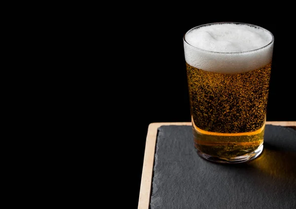 Glass of lager beer with foam and bubbles on stone board on black background. Space for text — Stock Photo, Image