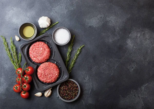 Plateau en plastique avec burgers de bœuf maison hachés crus avec des épices et des herbes. Vue du dessus et espace pour le texte sur fond de table de cuisine en pierre avec tomates sel et poivre . — Photo