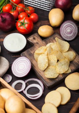 Taze organik ev yapımı patates ekşi krema ve koyu ahşap arka plan üzerinde doğrama tahtası üzerinde kırmızı soğan ile cips cips. Yeşil soğan ve ketçap kase ile Domates.