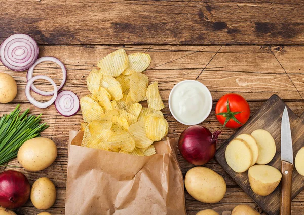 Frites de pommes de terre fraîches bio maison croustilles avec crème sure et oignons rouges et épices dans un sac en papier sur fond en bois. Avec pommes de terre jaunes fraîches — Photo