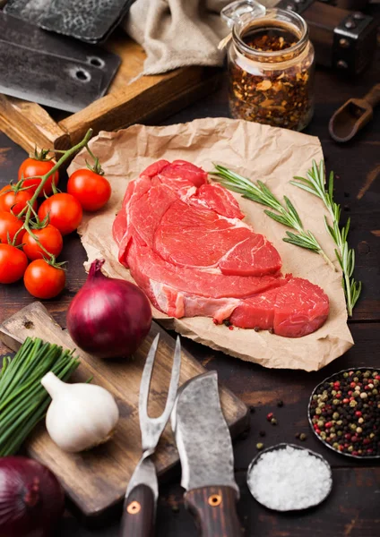 Fresh raw organic slice of braising steak fillet on butchers paper with fork and knife on dark wooden background. Red onion, tomatoes with salt and pepper. — Stock Photo, Image