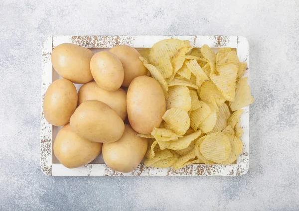 Fresh organic homemade potato crisps chips with raw yellow potatoes in white wooden box on light kitchen table background. — Stock Photo, Image