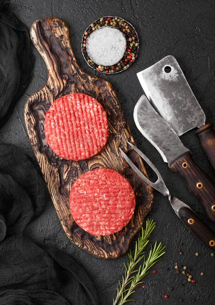 Los agricultores caseros picados crudos frescos asan hamburguesas de carne de res en la tabla de cortar vintage con especias y hierbas y hacha de carne, tenedor y cuchillo en la tabla negra . —  Fotos de Stock