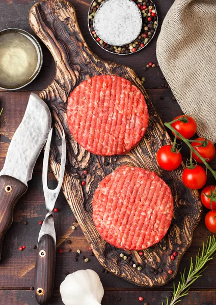 Verse rauwe gehakte zelfgemaakte boeren Grill rundvlees hamburgers op vintage snijplank met kruiden en kruiden en vork met mes op houten plank. Olie, knoflook en tomaten. — Stockfoto