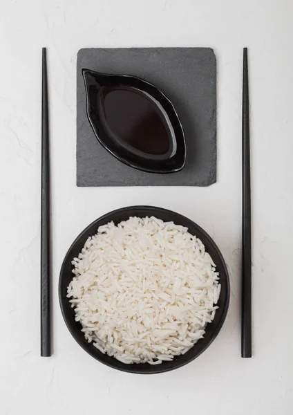 Tigela preta com arroz de jasmim de basmati orgânico cozido com pauzinhos pretos e molho de soja doce no tapete de pedra no fundo branco . — Fotografia de Stock