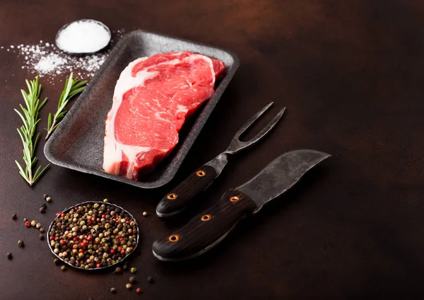 Raw sirloin beef steak in plastic tray with knife and fork on rusty background. Salt and pepper with fresh rosemary — Stock Photo, Image