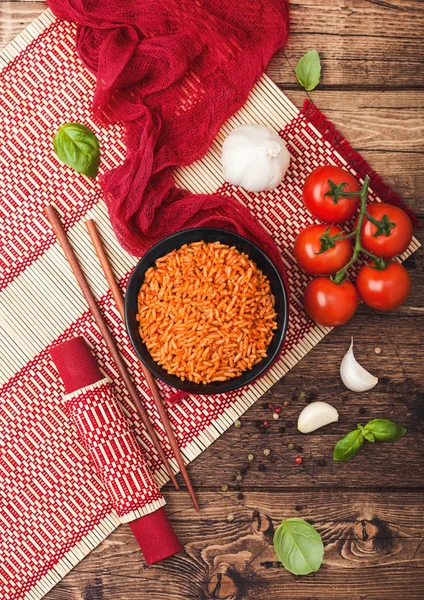 Black plate bowl of rice with tomato and basil and garlic and chopsticks on red bamboo place mat on wooden background with red cloth. Top view. — Stock Photo, Image