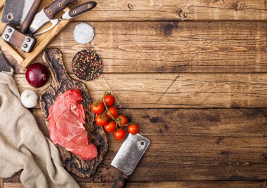 Fresh raw organic slice of braising steak fillet on chopping board with meat hatchet on wooden background. Red onion, tomatoes with salt and pepper. Space for text clipart