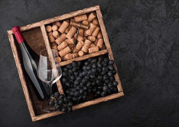 Bottle of red wine and empty glass with dark grapes with corks and corkscrew inside vintage wooden box on black stone background. Top view — Stock Photo, Image