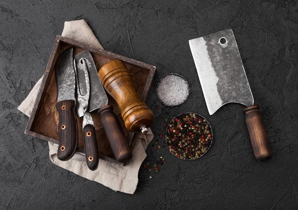 Cuchillo de carne vintage y tenedor y hacha en caja de madera vieja sobre fondo de mesa negro. Utensilios de carnicero. Sal y pimienta — Foto de Stock