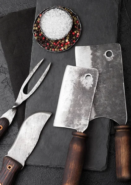 Coltello e forchetta da carne vintage e accette con tagliere in pietra e fondo tavola nero. Utensili da macellaio. Da vicino. — Foto Stock