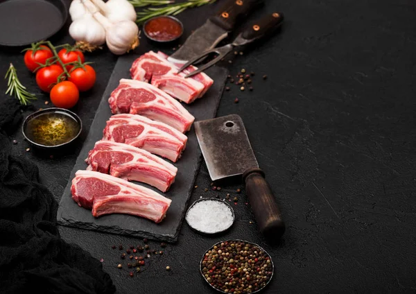 Fresh raw butchers lamb beef cutlets on stone board with vintage meat fork and knife and hatchet on black background.Salt, pepper and oil with tomatoes and garlic and barbeque sauce.Space for text — Stock Photo, Image
