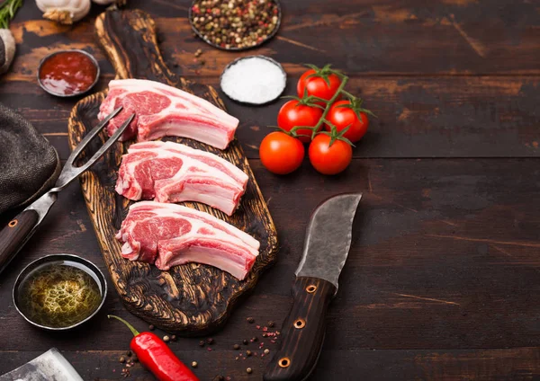 Fresh raw butchers lamb beef cutlets on chopping board with vintage meat fork and knife on wooden background.Salt, pepper and oil with tomatoes and garlic and barbeque sauce. — Stock Photo, Image
