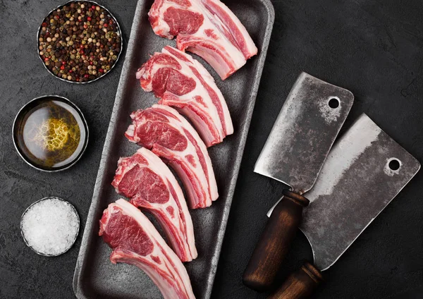 Verse rauwe slagers lamsvlees koteletten in kunststof tray met vintage vlees hakmessen op zwarte achtergrond. Zout, peper en olie in stalen kom. — Stockfoto