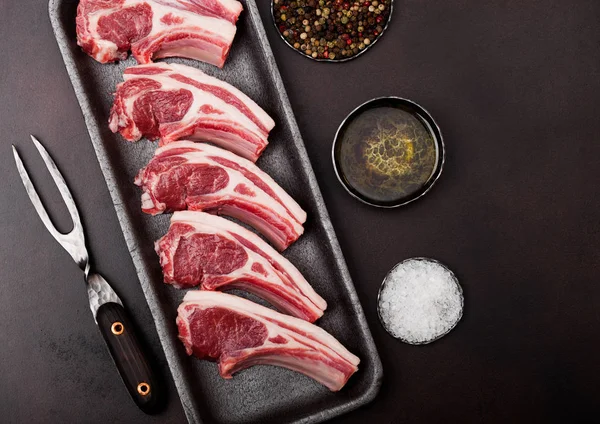 Verse rauwe slagers lamsvlees koteletten in kunststof tray met vintage Vleesvork op bruine achtergrond. Zout, peper en olie in stalen kom. — Stockfoto