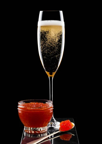 Elegante copa de champán amarillo con caviar rojo sobre cuchara dorada y recipiente de vidrio de caviar sobre tabla de mármol sobre fondo negro . — Foto de Stock