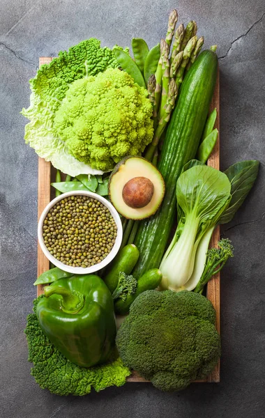 Assorted green toned raw organic vegetables in wooden box on dark background. Avocado, cabbage, cauliflower and cucumber with trimmed and mung beans and pepper and broccoli with sparagus tips. — Stock Photo, Image