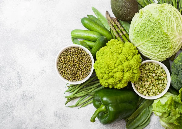 Assorted green toned raw organic vegetables on white stone background. Avocado, cabbage, broccoli, cauliflower and cucumber with trimmed mung beans and split peas in white bowl. — Stock Photo, Image