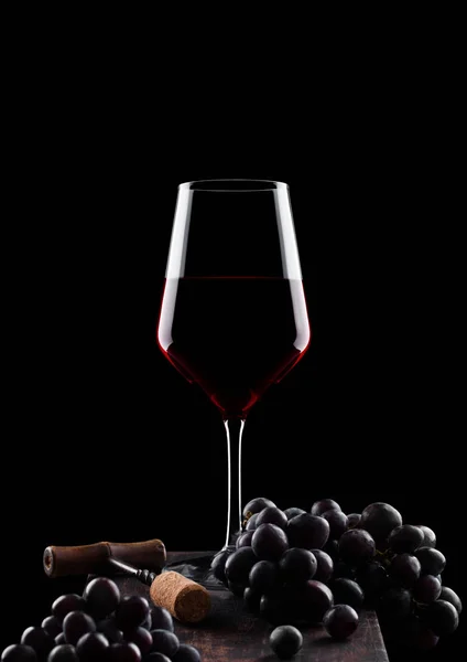 Glas rode wijn met donkere druiven en vintage kurkentrekker opener en kurk op houten bord op zwarte achtergrond. — Stockfoto