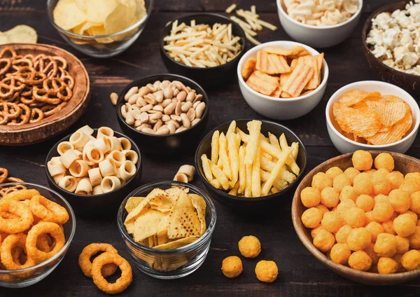 Todos los snacks clásicos de papa con cacahuetes, palomitas de maíz y aros de cebolla y pretzels salados en platos hondos sobre fondo negro. Giros con palitos y papas fritas y patatas fritas con nachos y bolas de queso . — Foto de Stock