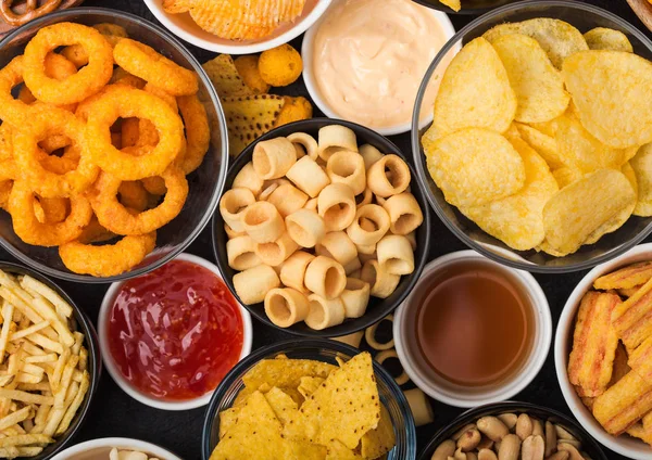 All classic potato snacks with peanuts, popcorn and onion rings and salted pretzels in bowl plates on black background. Twirls with sticks and potato chips and crisps with nachos and cheese balls. — Stock Photo, Image