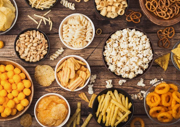 Tutti i classici snack di patate con arachidi, popcorn e anelli di cipolla e pretzel salati in piatti ciotola su sfondo di legno. Giri con bastoncini e patatine e patatine con nachos e palline di formaggio . — Foto Stock