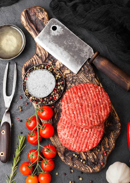 Los granjeros caseros picados crudos frescos asan hamburguesas de carne de res en la tabla de cortar vintage con especias y hierbas y hacha de carne en la tabla negra. Aceite, ajo y tomates . —  Fotos de Stock