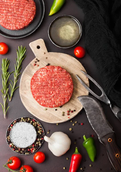 Los agricultores caseros picados crudos frescos asan hamburguesa de carne de res en la tabla de picar redonda con especias y hierbas y tenedor y cuchillo en la tabla marrón. Aceite, pimienta y ajo . —  Fotos de Stock