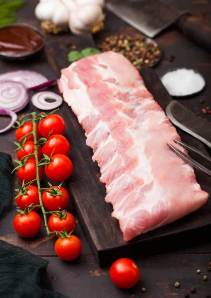 Côtes de porc crues fraîches sur planche à découper et fourchette à viande vintage et couteau sur fond en bois. Tomates fraîches et oignon rouge à l'ail, sel et poivre . — Photo